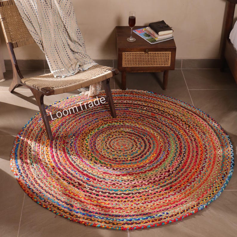 May include: A round, multi-colored jute rug with concentric circles of red, orange, blue, and green. The rug is on a tiled floor, with a wooden chair and side table in the background. The text "LoomTrade" is visible on the rug.