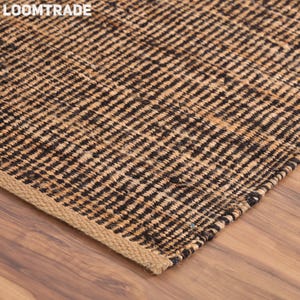 May include: A rectangular area rug featuring a woven texture with alternating vertical stripes of natural tan and black fibers. The rug has a visible braided edge finish and the text LOOMTRADE is printed in white in the upper left corner.