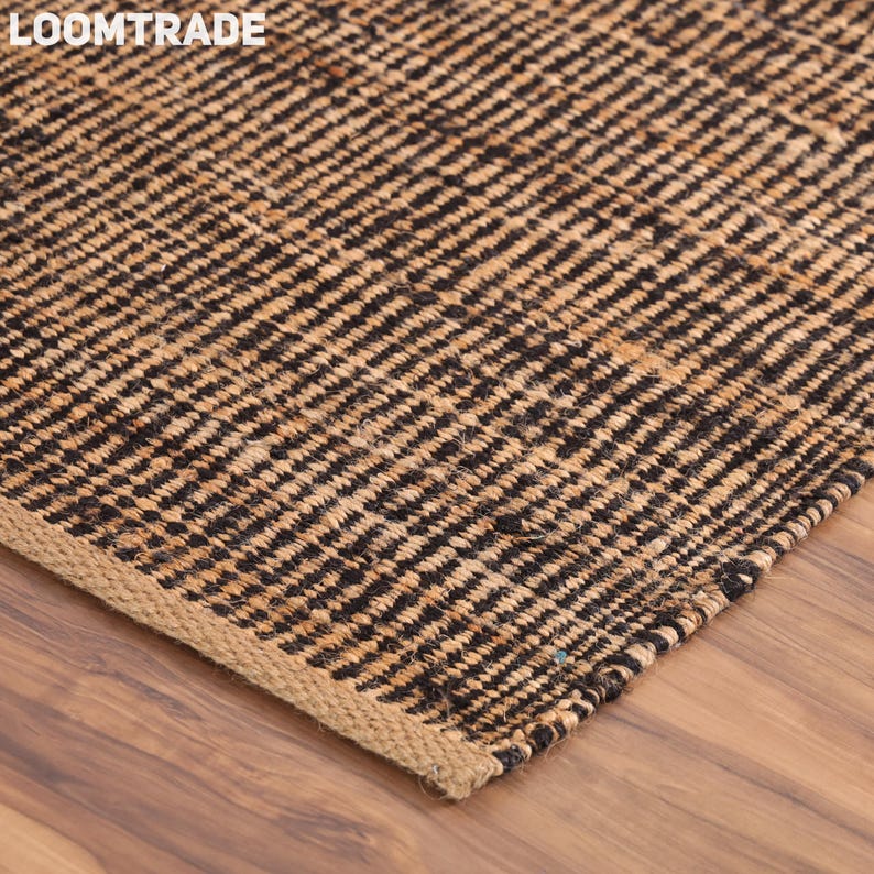 May include: A rectangular area rug featuring a woven texture with alternating vertical stripes of natural tan and black fibers. The rug has a visible braided edge finish and the text LOOMTRADE is printed in white in the upper left corner.