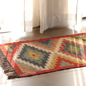 以下が含まれることがあります： 赤色の縁取りと、グレー、黄色、オレンジ、青色の幾何学模様が特徴の長方形のラグ。 ラグにはフリンジの縁があり、左下に「LoomTrade」の文字が見えます。 廊下やリビングスペースに適しています。