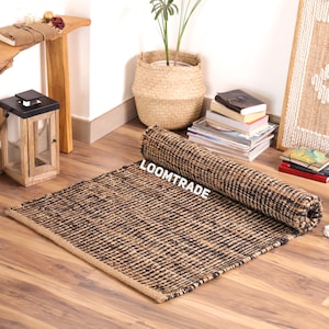 May include: A rectangular area rug featuring a woven texture with alternating black and natural tan fibers. The rug is partially rolled up at one end and displays the white text LOOMTRADE printed clearly across the center of the woven surface.
