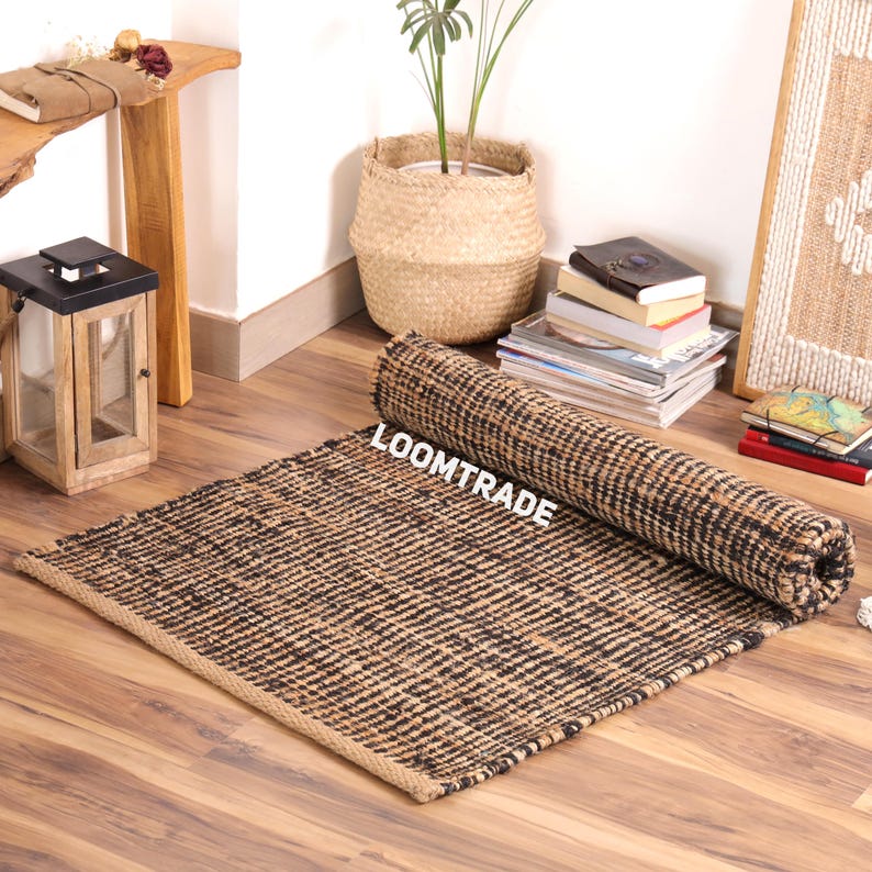 May include: A rectangular area rug featuring a woven texture with alternating black and natural tan fibers. The rug is partially rolled up at one end and displays the white text LOOMTRADE printed clearly across the center of the woven surface.