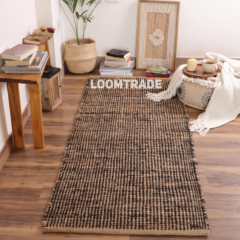 May include: A rectangular area rug featuring a woven texture with alternating vertical stripes of natural tan and black fibers. The rug has a consistent flat-weave construction and is positioned centrally, displaying the text LOOMTRADE in white.