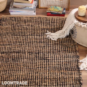 May include: A rectangular area rug featuring a woven pattern of alternating natural tan and black vertical stripes. The rug has a textured, coarse surface appearance and displays the white text LOOMTRADE in the bottom left corner.