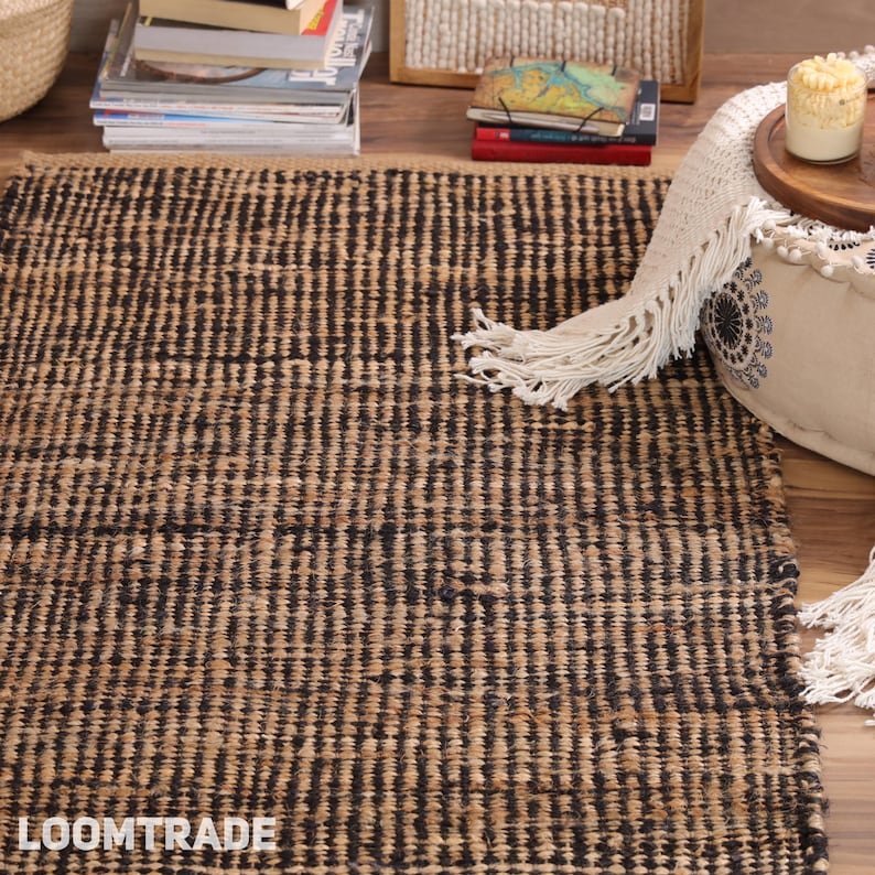 May include: A rectangular area rug featuring a woven pattern of alternating natural tan and black vertical stripes. The rug has a textured, coarse surface appearance and displays the white text LOOMTRADE in the bottom left corner.