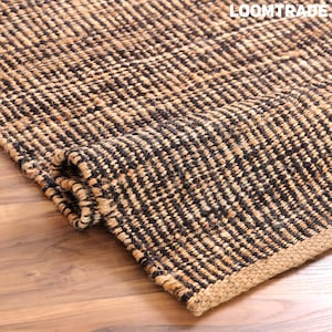 May include: A woven area rug featuring a textured pattern of alternating black and natural tan fibers. The rug is partially folded, revealing its thick, coarse construction and a solid tan border. The text LOOMTRADE is visible in the corner.