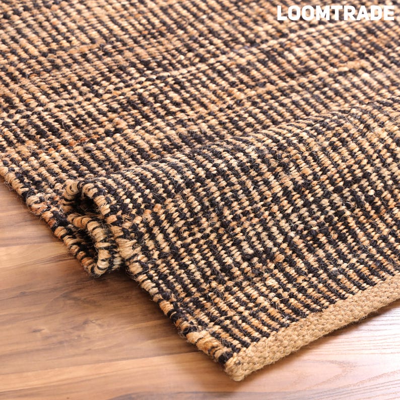 May include: A woven area rug featuring a textured pattern of alternating black and natural tan fibers. The rug is partially folded, revealing its thick, coarse construction and a solid tan border. The text LOOMTRADE is visible in the corner.