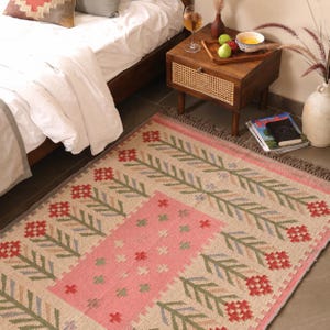 May include: A rectangular woven area rug featuring a central pink panel with small cross motifs, surrounded by a repeating pattern of red floral blooms and green leafy stems on a light beige background, finished with a fringed edge.