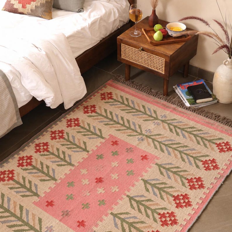 May include: A rectangular woven area rug featuring a central pink panel with small cross motifs, surrounded by a repeating pattern of red floral blooms and green leafy stems on a light beige background, finished with a fringed edge.