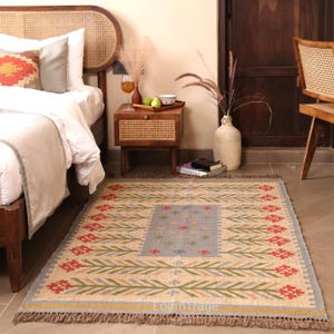 May include: A rectangular area rug featuring a beige base with a central light blue panel and a repeating floral pattern in red and green. The rug has a fringed edge and includes the text Loom Trade printed in white near the bottom border.