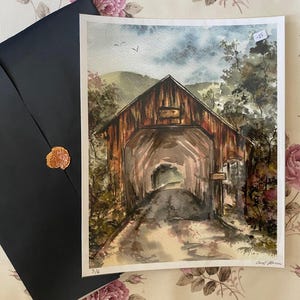 Op de afbeelding: Aquarel schilderij van een overdekte brug met een donkere, verweerde houten buitenkant. Het kunstwerk toont een weg die door de brug leidt, omgeven door bomen en een bewolkte lucht. Een zwarte envelop met een gouden zegel ligt naast het schilderij.
