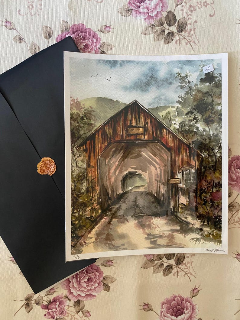 Op de afbeelding: Aquarel schilderij van een overdekte brug met een donkere, verweerde houten buitenkant. Het kunstwerk toont een weg die door de brug leidt, omgeven door bomen en een bewolkte lucht. Een zwarte envelop met een gouden zegel ligt naast het schilderij.
