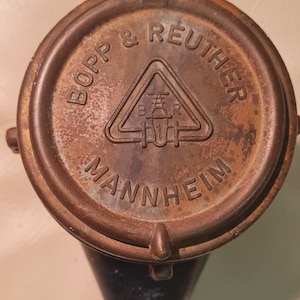 Può includere: Un oggetto metallico vintage color bronzo con la scritta "BOPP & REUTHER MANNHEIM" che circonda un emblema centrale. L'emblema è un triangolo con le lettere "B R". L'oggetto ha una base grigio scuro e una parte superiore grigio chiaro.