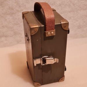 Puede incluir: Caja rectangular verde oliva con asa de cuero marrón y protectores de esquina de latón. La caja tiene un cierre de cierre metálico. El asa está sujeta con dos remaches de latón a cada lado. Estilo vintage.