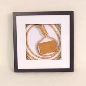 May include: A framed artwork featuring a coiled rope with a noose and a rectangular, tan-colored object suspended within. The artwork is encased in a black frame with a white matting, creating a minimalist aesthetic.