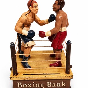 Könnte beinhalten: Eine mechanische Sparbüchse aus Gusseisen mit zwei Boxern in einem Ring. Ein Boxer trägt blaue Shorts und Handschuhe, der andere rote Shorts und Handschuhe. Der Sockel ist gelb mit braunem Rand und der Aufschrift "Boxing Bank" in weißen Buchstaben.