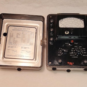 May include: Vintage black and silver electrical testing device with an open lid revealing a detailed diagram. The device features a gauge, dials, and various connection points. The open lid displays a detailed chart.