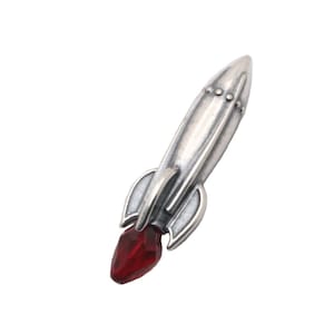 May include: A silver rocket ship charm with a red crystal at the tip. The rocket has a detailed design and a vintage look.