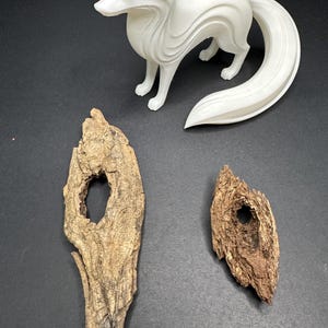 Könnte beinhalten: Eine weiße, 3D-gedruckte Fuchsfigur mit gebogenem Schwanz steht über zwei Treibholzstücken. Die Treibholzstücke sind braun mit Löchern und strukturierten Oberflächen. Der Hintergrund ist schwarz.
