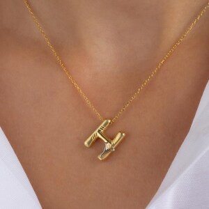 May include: A gold necklace featuring an "H" pendant. The pendant has a textured finish and a small, clear detail. The necklace is worn over a white shirt.