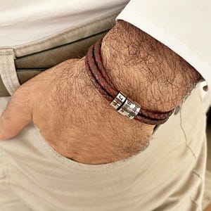 May include: Braided burgundy leather bracelet with three silver beads. Each bead has a name engraved on it. The bracelet is wrapped around a wrist, with the person's hand in a pocket.