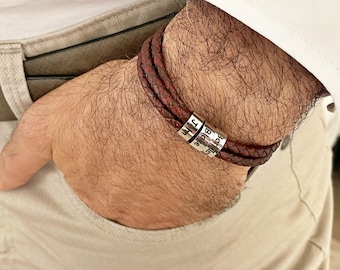 May include: Burgundy leather braided bracelet with three silver beads, each engraved with a name. The bracelet is wrapped around a wrist. The person's hand is in a pocket.