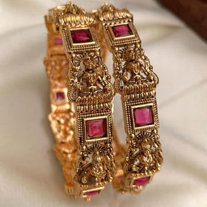 May include: Gold-toned bangles with intricate carvings and embedded red gemstones. The bangles feature detailed figures and square-cut stones, showcasing a traditional design. The jewelry is displayed on a soft, neutral background.