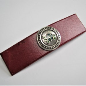 May include: A burgundy leather hair clip with a silver circular design featuring a white and green enamel flower.