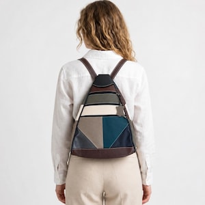Op de afbeelding: Een driehoekige rugzak met een color-block ontwerp in de kleuren zwart, bruin, crème, olijfgroen en blauwgroen. De tas heeft dunne bruine schouderbanden, een handvat aan de bovenkant en een klein leren label aan de ritssluiting.