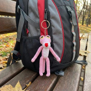 Può includere: Un portachiavi a forma di pantera rosa all'uncinetto, con lingua rossa e occhi gialli, è appeso a uno zaino. La pantera fatta a mano ha braccia e gambe lunghe e una pancia bianca. Lo zaino è nero, rosso e grigio.