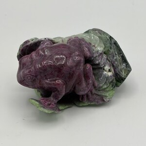 May include: A carved stone frog sculpture in shades of deep red and green. The frog is detailed with visible eyes and textured skin, resting on a bed of carved leaves. The sculpture is a decorative item.