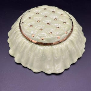 May include: A pale green ceramic flower frog with a ruffled edge. The top has a grid of holes and is trimmed with a gold band and gold accents. The flower frog is designed to hold flowers in place.