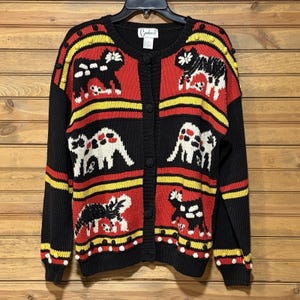 Può includere: Un cardigan nero, rosso e giallo con chiusura a bottoni. Il maglione presenta un motivo ripetuto di figure nere e bianche simili a mucche su pannelli rossi, con strisce gialle e dettagli di pompon bianchi.