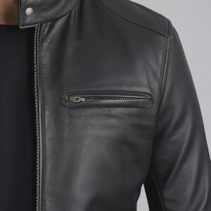 May include: A close-up of a black leather jacket. The jacket has a stand-up collar with a snap closure, a zippered chest pocket, and a full-length zipper. The jacket's leather appears smooth and has a slight sheen.