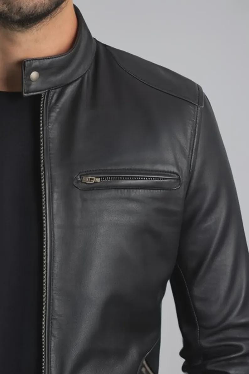 May include: A close-up of a black leather jacket. The jacket has a stand-up collar with a snap closure, a zippered chest pocket, and a full-length zipper. The jacket's leather appears smooth and has a slight sheen.