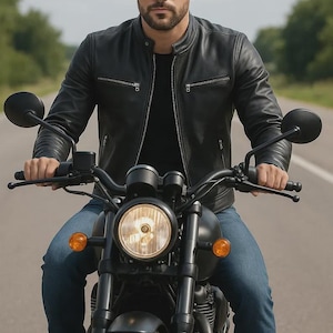 May include: A person wearing a black leather jacket and blue jeans rides a black motorcycle on a paved road. The motorcycle has a round headlight and orange turn signals. The background is blurred with green trees.
