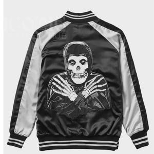 May include: Black satin bomber jacket with a white and silver design. The back features a large graphic of a skull with crossed skeletal hands. The jacket has white stripes on the cuffs, collar, and hem.