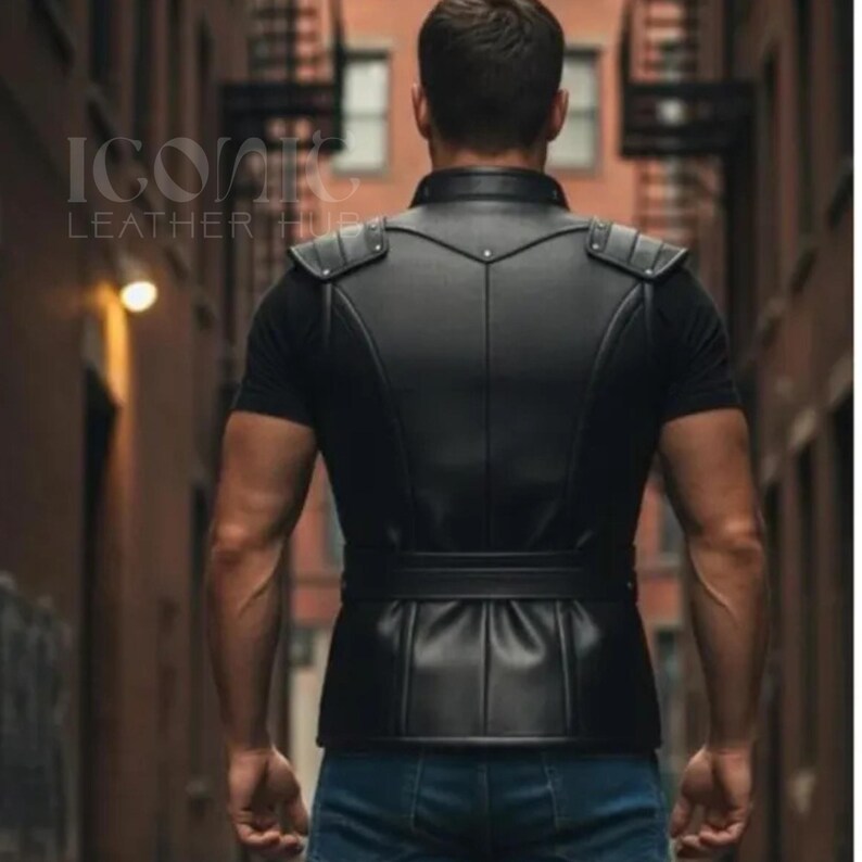 May include: A black leather vest with shoulder accents and a belted waist. The vest is worn over a black t-shirt. The person is wearing blue jeans. The image is taken from behind, in an alleyway.