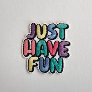 Könnte beinhalten: Ein farbenfroher, gestickter Aufnäher mit den Worten "JUST HAVE FUN" in Buchstaben. Der Text ist in Lila-, Türkis-, Rosa-, Gelb- und Blautönen gehalten und schwarz umrandet. Der Aufnäher befindet sich auf weißem Hintergrund.