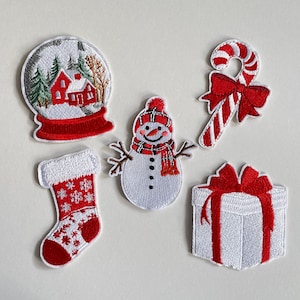 Könnte beinhalten: Gestickte Weihnachtspatches mit Schneekugel, Schneemann, Zuckerstange, Strumpf und Geschenkbox. Die Patches sind weiß mit roten Akzenten und Details.