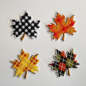 Könnte beinhalten: Vier gestickte Ahornblatt-Aufnäher in Herbstfarben. Ein Blatt ist schwarz-weiß kariert, ein anderes orange und rot, ein drittes gelb und orange kariert und das letzte grün, rot und weiß kariert.