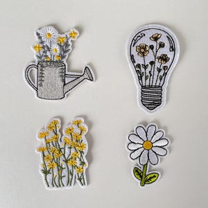 Puede incluir: Cuatro parches bordados con diseños florales. Uno representa una regadera llena de margaritas. Otro muestra una bombilla con flores en su interior. Los otros dos parches son de flores amarillas y una margarita.