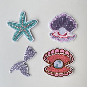 Könnte beinhalten: Vier gestickte Aufnäher mit Meerjungfrauen-Motiv. Enthält einen türkisfarbenen Seestern mit rosa Akzenten, eine lila Meerjungfrauenschwanzflosse und zwei offene Muscheln mit Perlen. Die Aufnäher haben weiße Ränder.