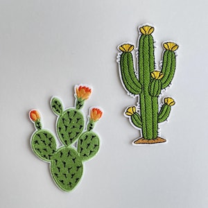 以下が含まれることがあります： 2つの刺繍されたサボテンのパッチ。1つのパッチは、黄色の花と茶色のベースを持つ緑色のサボテンを示しています。もう1つのパッチは、オレンジ色の花を持つ緑色のサボテンを示しています。両方のパッチには白い縁があり、白い背景にあります。