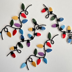 Puede incluir: Parches bordados de luces navideñas de colores. Cada bombilla es de un color diferente, incluyendo rojo, amarillo, verde, azul y morado. Las luces están conectadas por un cable negro, creando un diseño festivo.