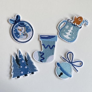 Könnte beinhalten: Eine Sammlung von gestickten Aufnähern mit winterlichem Feiertags-Thema. Die Aufnäher zeigen einen Schneemann in einer Schneekugel, einen Weihnachtsstrumpf, eine Tasse mit einem Lebkuchenmann, Weihnachtsbäume und eine Glocke mit einer Schleife. Die Aufnäher sind hauptsächlich blau und weiß.
