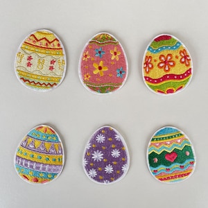 Könnte beinhalten: Sechs gestickte Ostereier-Patches mit farbenfrohen Designs. Die Eier zeigen Muster aus Blumen, geometrischen Formen und Herzen in verschiedenen Farben, darunter Gelb, Rosa, Lila und Blau, mit weißen Rändern.