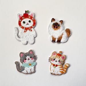 Könnte beinhalten: Vier gestickte Katzenaufnäher in verschiedenen Designs. Eine weiße Katze trägt einen roten Apfelhut. Eine andere ist eine flauschige braun-weiße Katze. Eine grau-weiße Katze hat einen blauen Kragen, und eine orangefarbene Tabby-Katze trägt einen Hut.