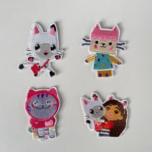 May include: Four embroidered patches depicting cartoon cat characters. The patches feature different designs, including a cat with a red bag, a cat in a pink hat, and a cat hugging a person. The patches are colourful and detailed.