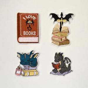 Peut inclure: Quatre écussons brodés représentant des dragons et des livres. L'un des écussons porte l'inscription "I LOVE BOOKS" avec une illustration de dragon. D'autres écussons montrent des dragons avec des livres. Les écussons sont dans les tons de marron, bleu, noir et blanc.